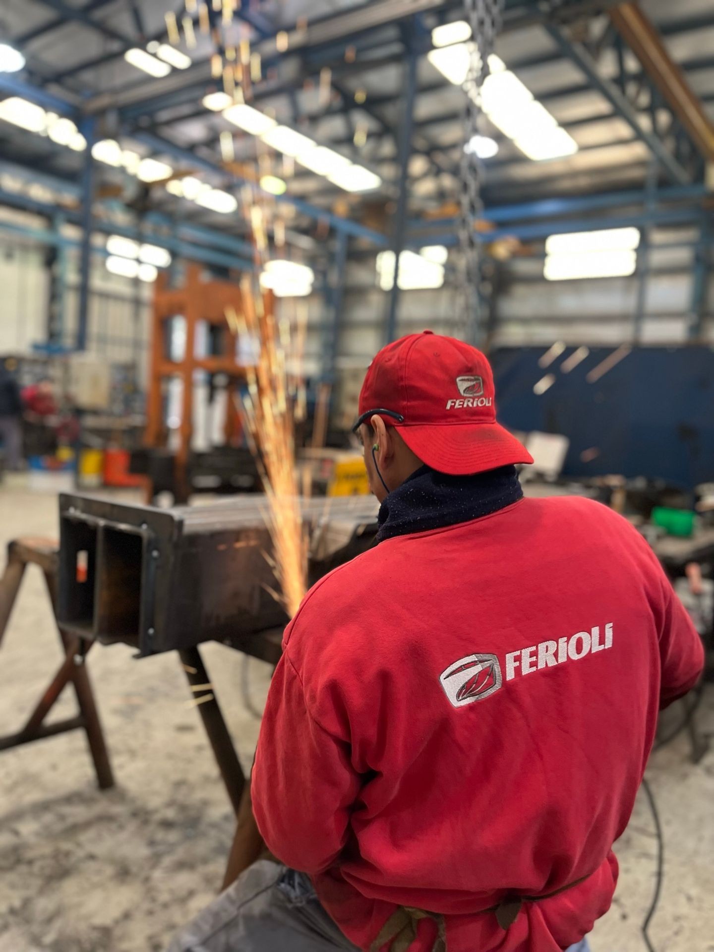 Worker in red jacket and cap using a grinder, creating sparks in a metal workshop.